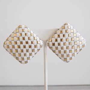 Op de afbeelding: Een paar vierkante oorbellen van wit en goud geweven materiaal. De oorbellen zijn gemaakt van een geweven materiaal met een geruit patroon.