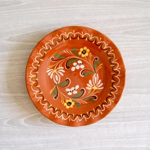 May include: A round terracotta-colored ceramic plate featuring a hand-painted floral motif in white, yellow, and green. The rim is decorated with a repeating black and yellow wavy line pattern, while the center displays stylized flowers and leaves.