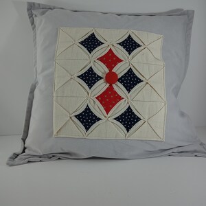 May include: A grey pillow with a white, red and blue quilted design. The design features a central red button and a pattern of diamonds and stars.
