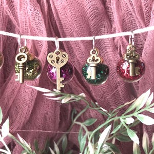 May include: Six glass ornaments with gold key charms and glitter inside. The ornaments are hanging on a white string and are against a pink background.
