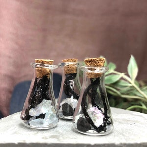 May include: Three small glass bottles with cork tops filled with black lava rock, iridescent crystals, and black string. The bottles are sitting on a gray surface.