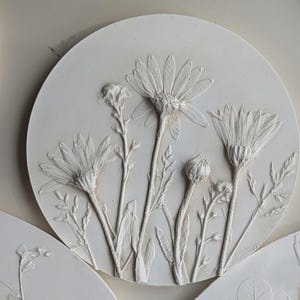 May include: Circular white wall art with a raised relief of daisy-like flowers and foliage. The flowers are in different stages of bloom, with detailed petals and stems. The artwork is a neutral colour.