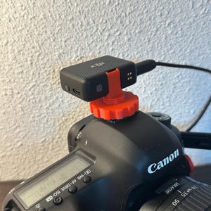 Pode incluir: Uma câmera Canon preta com uma lente e um pequeno receptor de áudio preto preso com uma montagem laranja. O receptor está conectado à câmera com um cabo preto. A câmera possui um visor digital e vários botões.