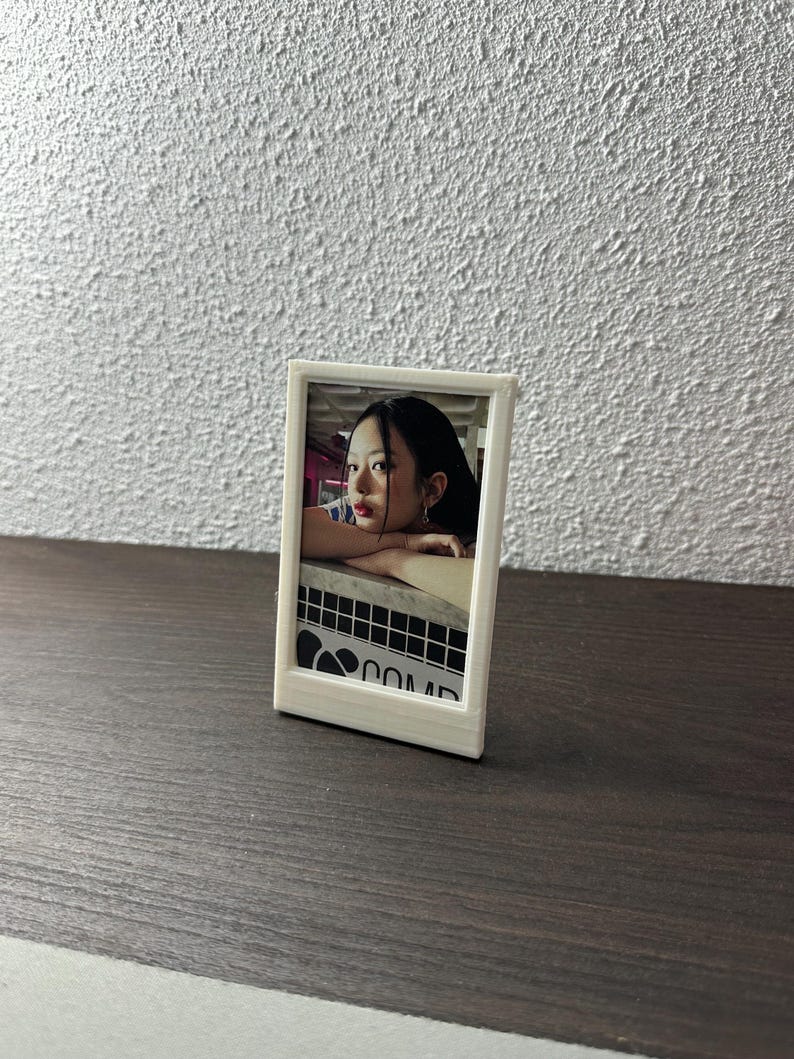 Peut inclure: Un cadre photo blanc avec une photo d'une femme &agrave; l'int&eacute;rieur. La photo montre une femme aux cheveux fonc&eacute;s portant une chemise blanche. Le cadre est pos&eacute; sur une surface en bois.