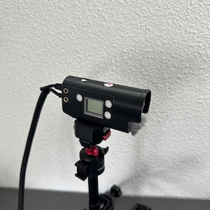 May include: A black camera with a small microphone attached to the top, mounted on a black tripod with a red knob. The camera has a small LCD screen and several buttons.