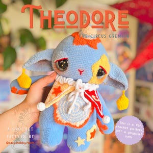 Puede incluir: Una criatura de ganchillo azul con grandes ojos oscuros, cabeza puntiaguda y orejas grandes. Lleva un cuello de volantes blanco con detalles triangulares naranjas y rojos. El texto dice THEODORE THE CIRCUS GREMLIN y A CROCHET PATTERN BY @cozy.hobby.room.