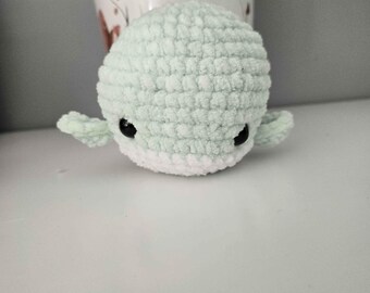 May include: A small, round, crocheted plush toy shaped like a whale. The body is made of soft, light mint green yarn with a white underside. It features two small side fins and two black, glossy, circular plastic eyes positioned on the front.