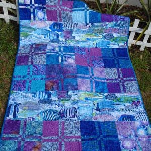 May include: A handmade quilt featuring a vibrant aquatic theme. The quilt showcases a patchwork design with squares of blue, purple, and white fabrics. The central panels display various fish illustrations. The quilt is bordered with a solid blue fabric.