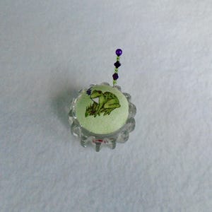 May include: A light green pincushion with a frog design, set in a clear, scalloped-edge holder. A beaded pin with purple beads extends upwards. The frog is green and white, with a small blue flower.