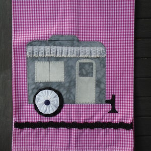 May include: A pink and white gingham tea towel with a gray and white appliqué of a camper van. The camper van has a white lace trim, a white wheel with a blue and white flower, and a black number "1" on the side.