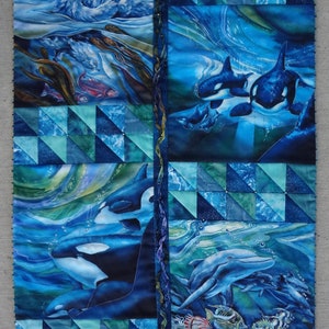 May include: A rectangular fabric wall hanging featuring a collage of marine life scenes in shades of blue, teal, and green. The quilt displays sea otters, orcas, and dolphins swimming in ocean waters, accented by triangular geometric border patterns.