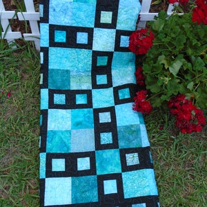 May include: A rectangular quilt featuring a patchwork design of various blue and teal fabric squares. The quilt is accented with a bold black grid pattern that separates the blue squares, and the entire surface displays a continuous meandering stitch.
