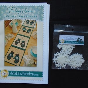 May include: A stack of paper with the text "The Vintage Series January Table Runner" and a picture of a table runner with mittens. Next to it is a bag of white felt snowflakes.