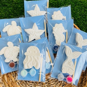 May include: A collection of paint-your-own sea creature kits, including starfish, seahorses, turtles, jellyfish, and mermaid tails. Each kit is individually packaged in a clear bag with a blue backing, paint pots, and a paintbrush. The kits are displayed in a woven basket.