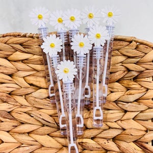 May include: A collection of clear plastic bubble wands, each topped with a white paper daisy featuring a yellow center. The wands are arranged in a cluster, with the white stems and circular bubble loops visible inside the transparent tubes.