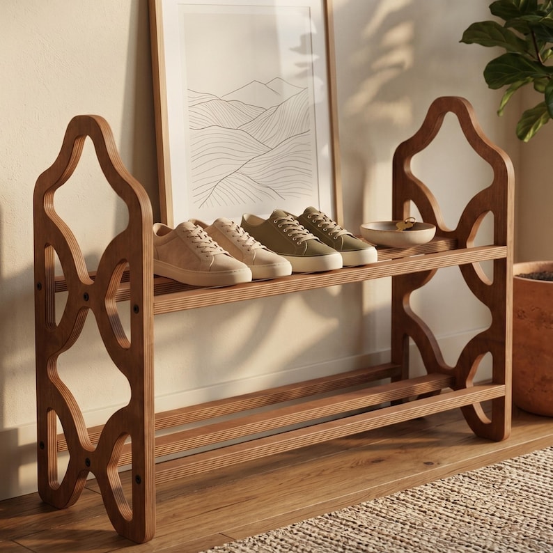 May include: A wooden shoe rack with two shelves, holding four pairs of sneakers in neutral colors. The rack has a decorative cutout design on the sides. A framed landscape art piece is in the background.