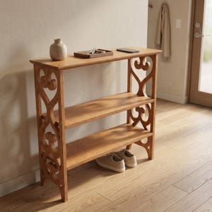 May include: A three-tiered wooden shelf with decorative side cutouts. The shelves hold a small vase, a tray with keys, and a phone. Two pairs of shoes are placed on the floor beneath the shelves. The wood has a warm, natural finish.