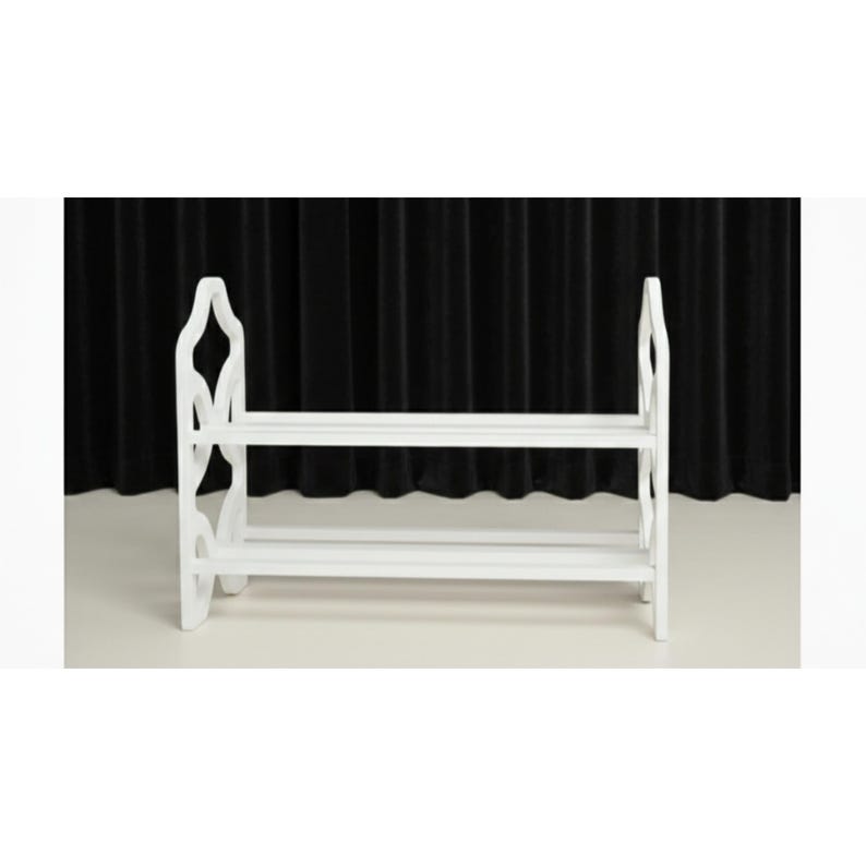 May include: A white wooden shoe rack with two shelves. The rack has decorative cutouts on the sides and is designed to hold multiple pairs of shoes. The background is a black curtain.