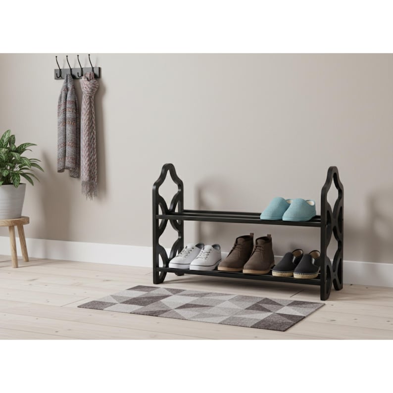 May include: A black two-tier shoe rack with a decorative design on the sides. The rack holds various shoes, including sneakers, boots, and slippers. A patterned rug sits beneath the rack. Scarves hang on a wall hook above the rack.