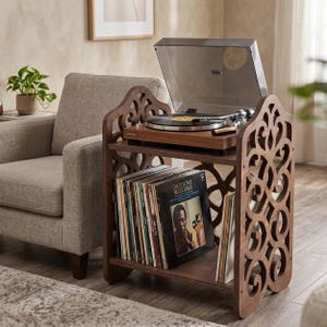 May include: A dark brown wooden record player stand with a turntable on top and a shelf filled with vinyl records. The stand has decorative cut-out designs on the sides. A gray armchair and a potted plant are in the background.