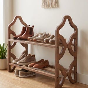 May include: A two-tiered, dark brown wooden shoe rack with an open, decorative design. The rack holds various pairs of shoes, including brown leather boots, sneakers, and flats. The rack is placed against a neutral wall.