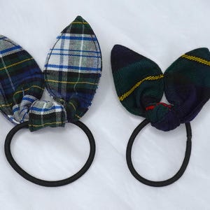 May include: Two plaid fabric hair ties with bunny ear-shaped bows. One tie features a blue, green, and white pattern, while the other has a green, navy, and yellow design. Both have black elastic bands.