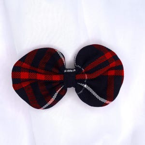 May include: A fabric hair bow featuring a dark navy blue and red plaid pattern with thin white accent lines. The bow is constructed with two rounded loops gathered at the center by a matching dark navy blue fabric band, creating a symmetrical shape.