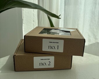May include: Two stacked, small brown cardboard boxes with clear window tops. Each box has a white label with the word "TEELICHTER" above the numbers "no. 1" and "no. 2" in a black, sans-serif font. The boxes are on a white surface.