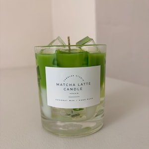 May include: A clear glass candle filled with green matcha latte colored wax and translucent ice cube shapes. A white rectangular label on the front reads CANDLES STUDIO MATCHA LATTE CANDLE VEGAN COCONUT WAX / HAND MADE with a single central wick.