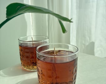 May include: Two clear glass candles with a textured pattern, filled with a brown liquid and a central wick. The candles sit on a white surface, with a green leaf and a white curtain in the background.