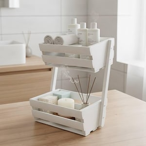 Könnte beinhalten: Ein weißes, zweistöckiges Holzregal mit Lamellendesign. Das obere Regal enthält gerollte Handtücher und Flaschen, während das untere Regal eine Kerze, Seife und Räucherstäbchen enthält. Das Regal steht auf einer Holzoberfläche.