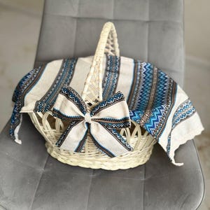 May include: A woven white basket with a handle, adorned with a matching bow and a patterned fabric. The fabric features a blue and white geometric design. The basket is sitting on a gray chair.