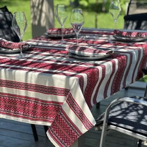 May include: A dining table set for an outdoor meal. The table is covered with a white and red striped tablecloth with a geometric pattern. Plates, napkins, and wine glasses are arranged on the table, ready for use.