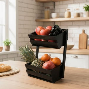 May include: A two-tiered black wooden fruit basket filled with fresh produce. The top tier holds red apples and black grapes, while the bottom tier contains a pineapple, oranges, and a pomegranate. The basket is on a wooden countertop.