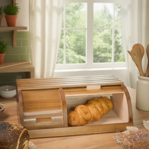 Könnte beinhalten: Eine Brotdose aus Holz mit Rolldeckel und Schiebetür. Die Dose ist aus hellem Holz gefertigt und enthält einen Laib geflochtenes Brot. Die Brotdose steht auf einem Holztisch in der Nähe eines Fensters.