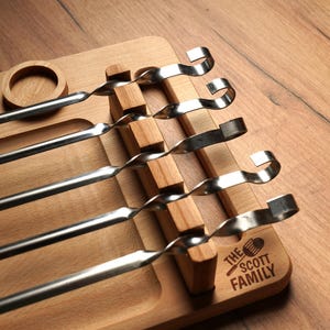 May include: A wooden serving board with a circular indentation and a rectangular section with wooden dividers. Six stainless steel skewers are placed in the dividers. The board is engraved with "THE SCOTT FAMILY" and a grill icon.