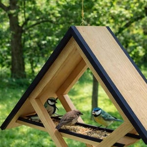 Hanging wooden bird feeder with a modern geometric design, featuring a triangular roof and open frame structure that allows birds to access seeds from multiple sides. The feeder is suspended by a cord and filled with birdseed, set against a blurred green garden background with trees and grass