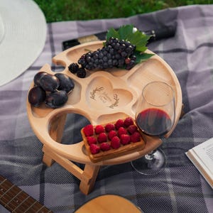 Könnte beinhalten: Ein helles Picknicktisch aus Holz mit runder Tischplatte und Klappbeinen. Der Tisch enthält ein Glas Rotwein, Trauben, Pflaumen, Brombeeren und eine Waffel mit Himbeeren. Der Tisch hat eine herzförmige Gravur mit den Namen "Johanna und David".