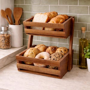 Op de afbeelding: Een houten broodmand met twee niveaus gevuld met diverse bakwaren, waaronder brood, bagels en muffins. De mand is gemaakt van donkerbruin hout en staat op een aanrecht. Een glazen pot, houten lepels en een fles olijfolie staan op de achtergrond.