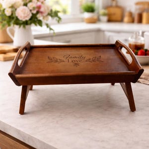 May include: A wooden serving tray with folding legs. The tray is a warm brown color and features handles on each end. The words "Family Love" are etched into the surface, surrounded by leaf designs. The tray is set on a marble countertop.