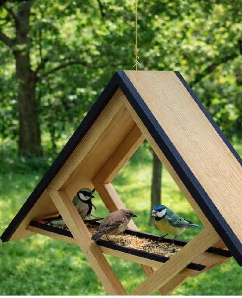 Hanging wooden bird feeder with a modern geometric design, featuring a triangular roof and open frame structure that allows birds to access seeds from multiple sides. The feeder is suspended by a cord and filled with birdseed, set against a blurred green garden background with trees and grass