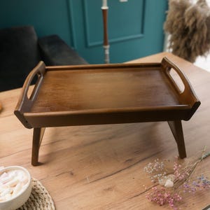 Könnte beinhalten: Ein braunes Holztablett mit klappbaren Beinen. Das Tablett hat zwei Griffe an den Seiten und ist perfekt zum Frühstück im Bett servieren.