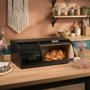 Puede incluir: Una panera de madera negra con tapa enrollable y panel frontal transparente. La panera es rectangular con un lado curvo y contiene varios panes. La panera está sobre una mesa de madera.
