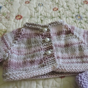 May include: A pink, white, and gray striped knitted cardigan with pearl buttons. The cardigan is on a white and floral patterned fabric.