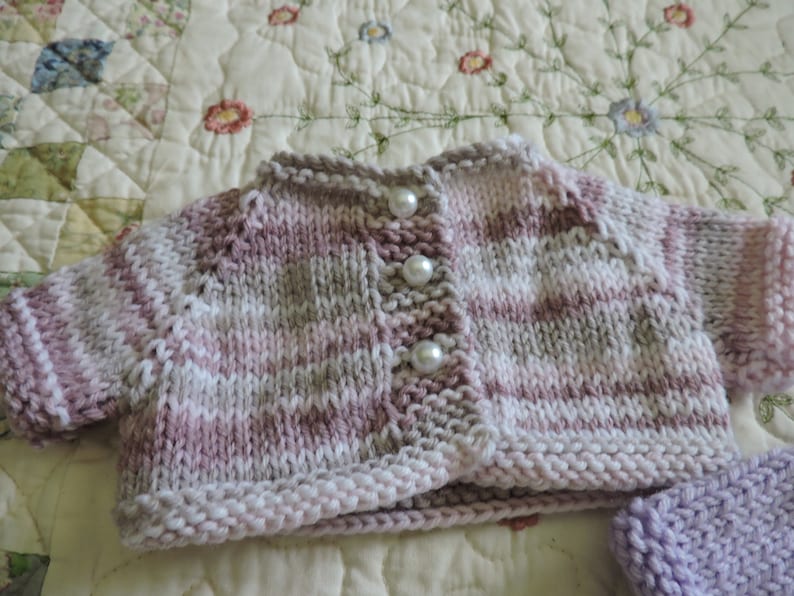 May include: A pink, white, and gray striped knitted cardigan with pearl buttons. The cardigan is on a white and floral patterned fabric.