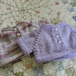 May include: Two knitted baby sweaters, one in a striped pattern of pink and white, and the other in a solid lavender color. Both sweaters have pearl buttons.