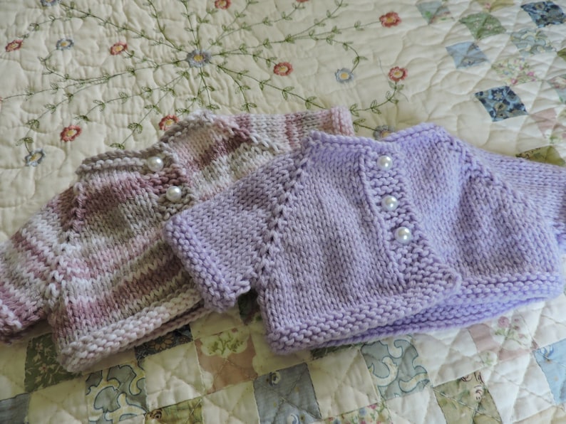 May include: Two knitted baby sweaters, one in a striped pattern of pink and white, and the other in a solid lavender color. Both sweaters have pearl buttons.