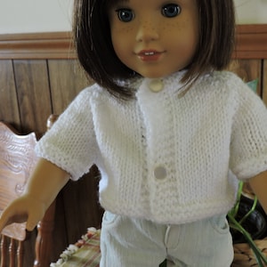 May include: A white knitted cardigan with a button closure. The cardigan is worn by a doll with brown hair and blue eyes.