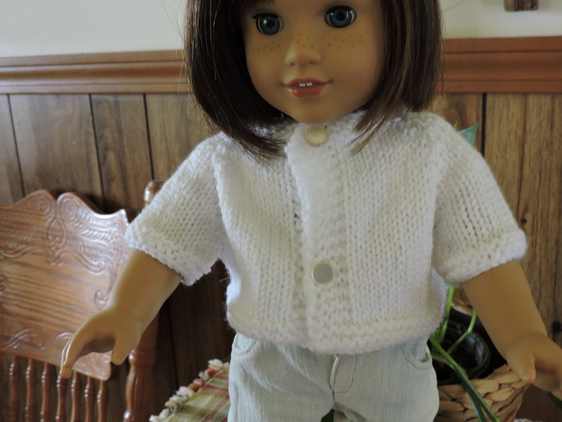 May include: A white knitted cardigan with a button closure. The cardigan is worn by a doll with brown hair and blue eyes.