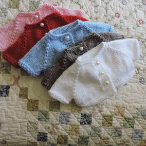 May include: Five knitted baby cardigans in different colors: pink, red, light blue, brown, and white. Each cardigan has pearl buttons.